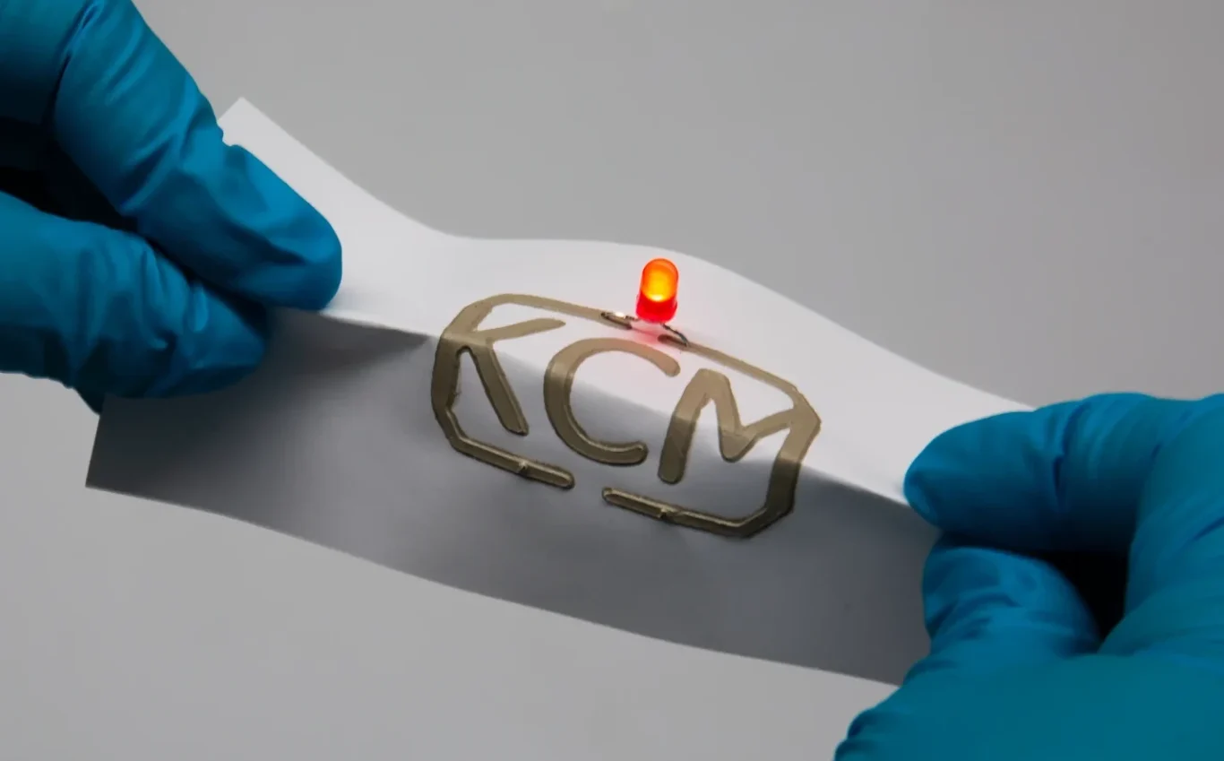 Flexible Electronics Demonstrator A person wearing blue gloves bends a sheet bearing the KCM logo and a red, glowing LED.